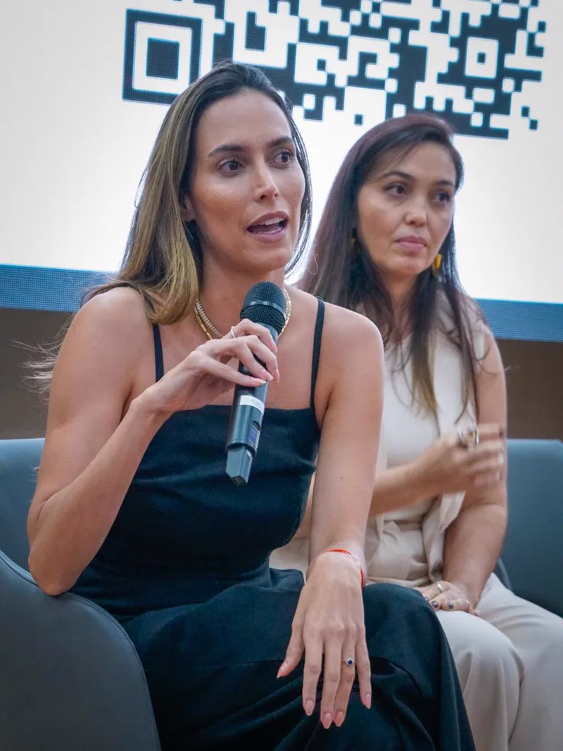 Marcella Teixeira respondendo perguntas com microfone em evento, ao lado de Milena Trindade